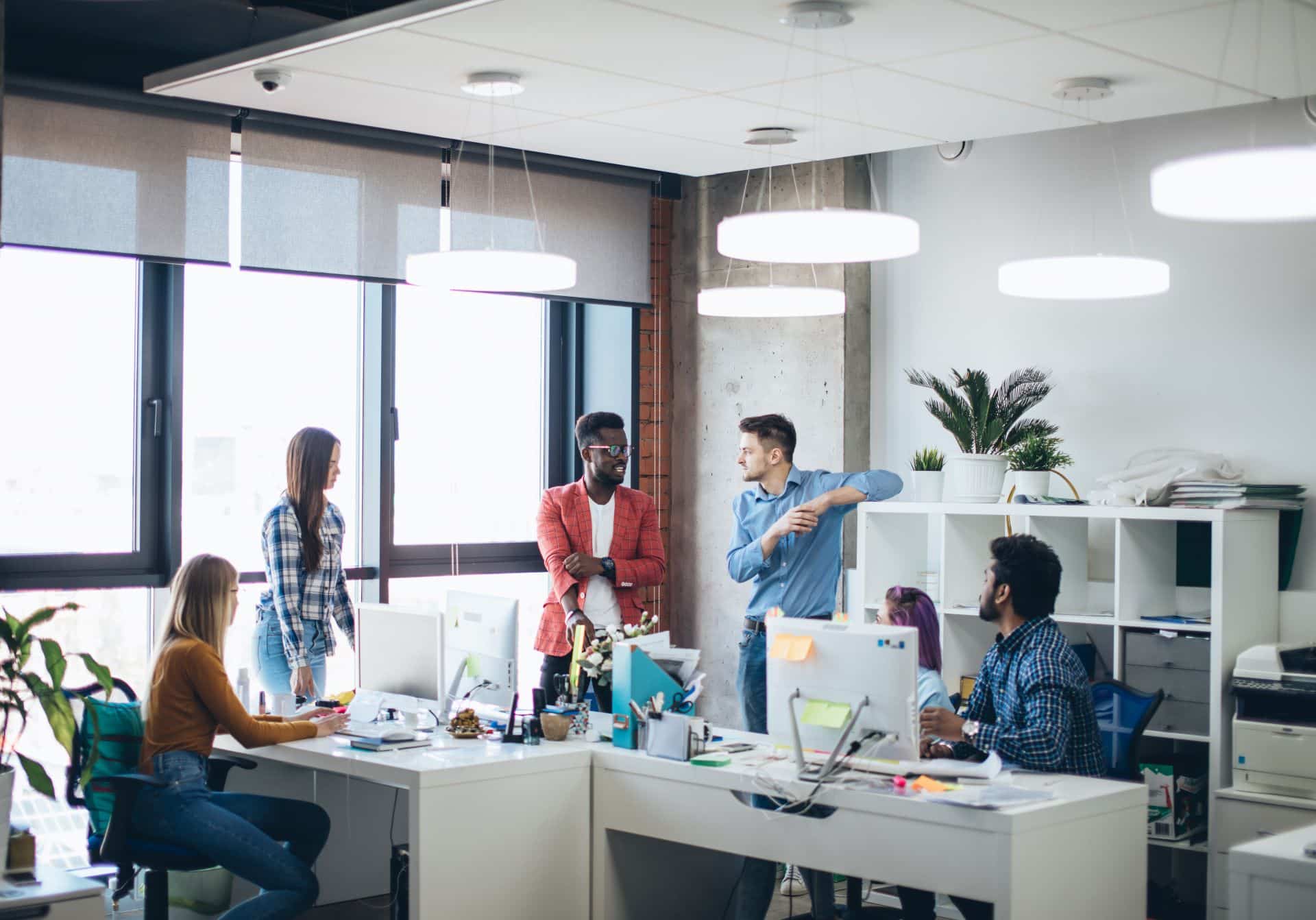 Team von Mitarbeitern im modernen Büro, das sich um Arbeitsplätze versammelt, um ein Projekt zu besprechen. Helle, offene Arbeitsumgebung mit großen Fenstern und minimalistischer Einrichtung.