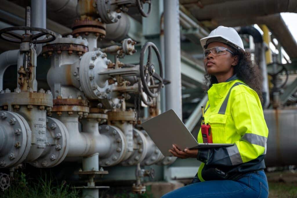 Eine Ingenieurin mit Laptop inspiziert eine Industrieanlage mit Rohrleitungen und Hydraulik Kupplung unter freiem Himmel
