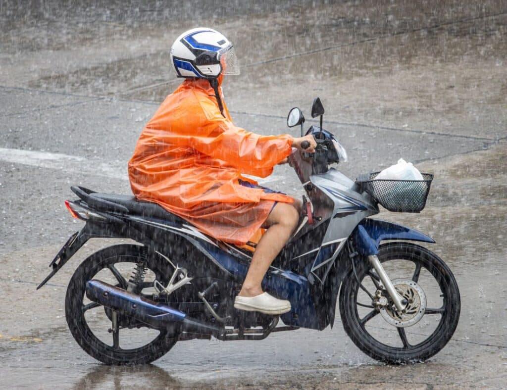 Person faehrt Motorroller im Regen, traegt orangefarbenen Regenponcho ohne Schutzbekleidung