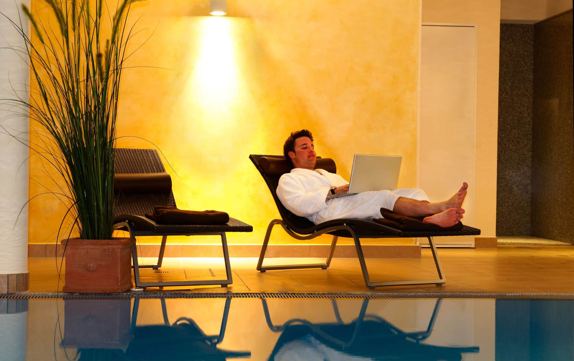 Mann im Bademantel mit Laptop auf Liege am Pool