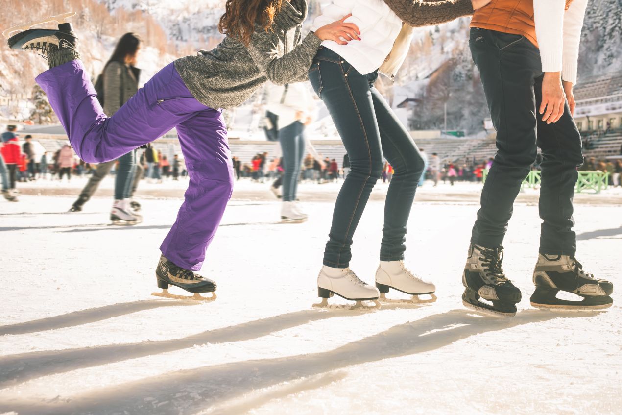 Drei Personen mit Schlittschuhen auf einer sonnigen Außen-Eisbahn – idealer Zeitpunkt für den Einsatz von Kufenschonern beim Gehen auf hartem Untergrund