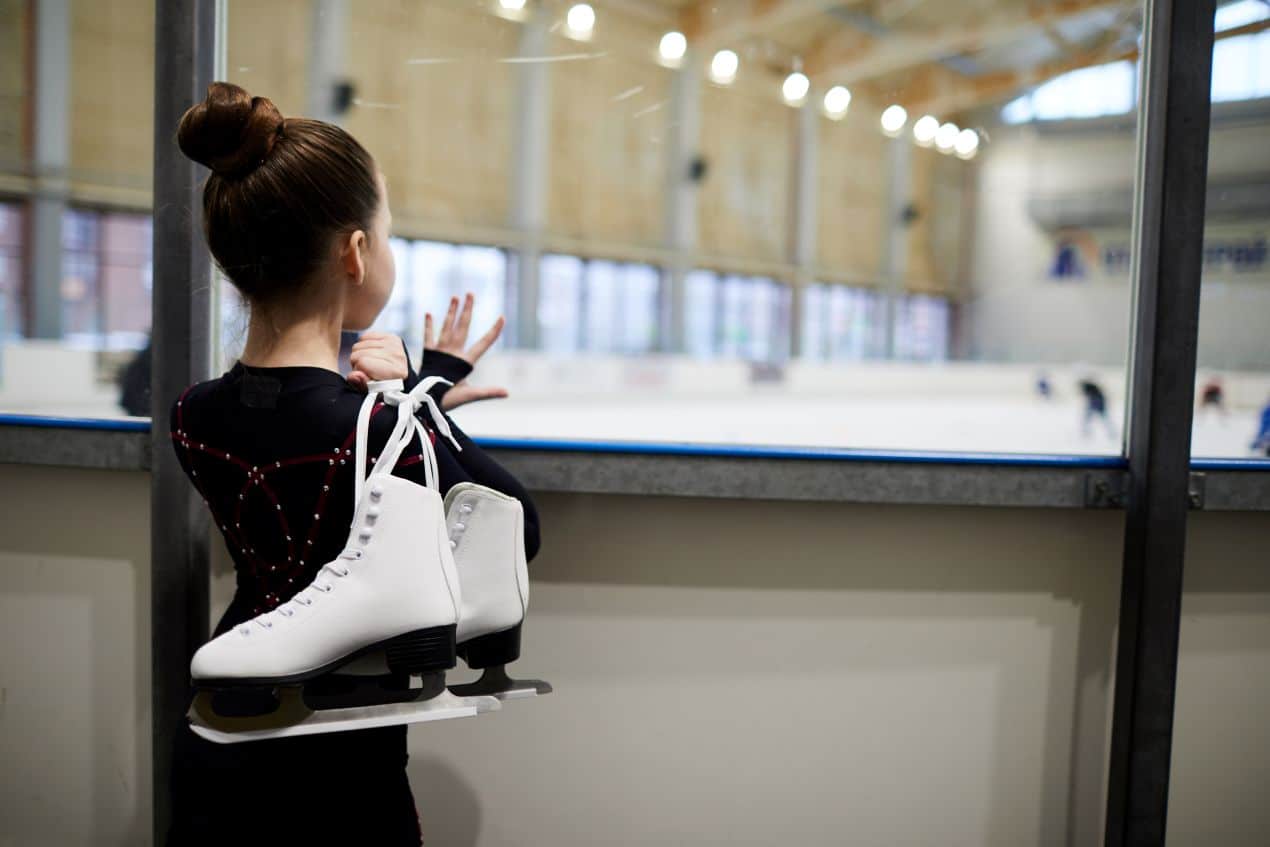 Junges Mädchen in Eiskunstlaufkleidung trägt weiße Schlittschuhe über der Schulter – perfekte Gelegenheit, Kufenschoner für sicheren Transport zu nutzen